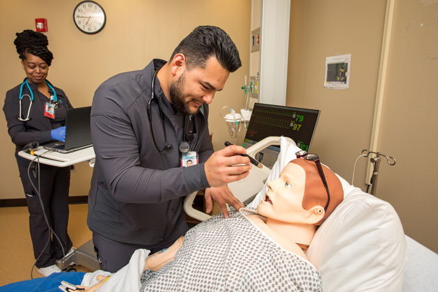 Two nursing students. One examining an patient mannequin while the other student is standing at a laptop.