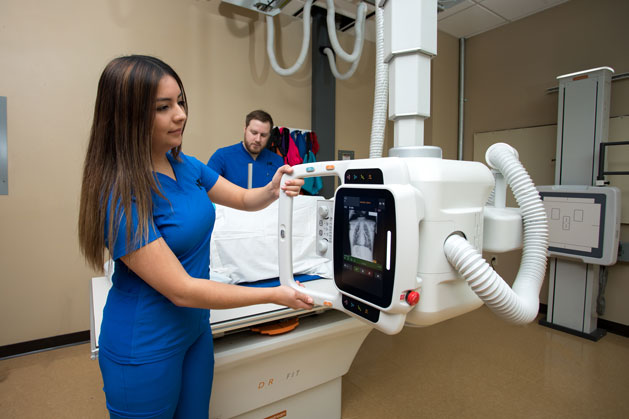 Two students in an x-ray classroom. One is standing by the xray table while one is pulling the x-ray machine down to line up the picture.