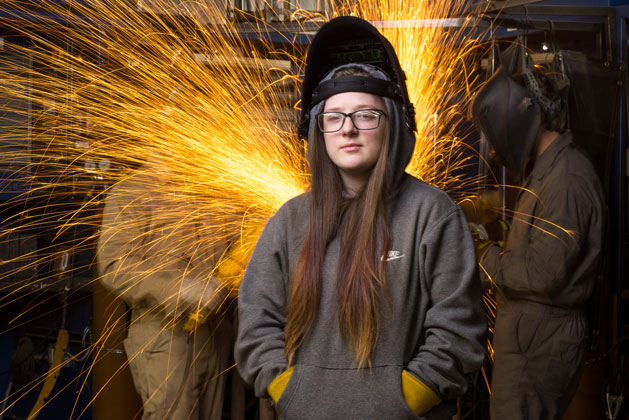 Female welder is facing the camera with a welding mask and gloves on while two students behind her make welding sparks fly.