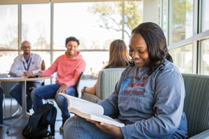 People sitting in a common area in a building. Several talking, one reading a textbook.