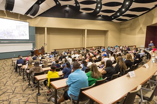 A wide-angle view from the back of the Martin Eisele Auditorium during a presentation. Attendees are seated at tiered, curved desks that face a large projection screen and a wooden podium. The room features a modern black and white acoustic ceiling treatment and neutral-toned walls.