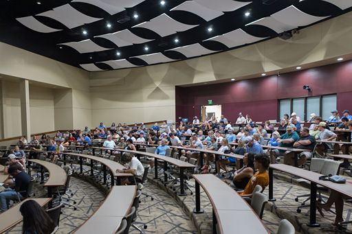 A side-view perspective of the Martin Eisele Auditorium showing the steep tiering of the curved seating rows. The image highlights the spacious desk workspace for each attendee and the integrated recessed lighting along the back wall, under a decorative acoustic ceiling.
