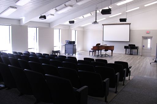 A perspective from the back of the Ellis Recital Hall looking toward the stage. The image highlights the 54 embedded theater seats, the open stage area with two pianos, and a large projection screen mounted on the far wall, demonstrating the room's suitability for both performances and presentations.