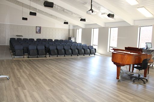A bright, wide-angle view of the Ellis Recital Hall stage from the side. The room features light wood-look flooring, large windows, and a grand piano positioned on the open stage area in front of several rows of dark upholstered theater seats.