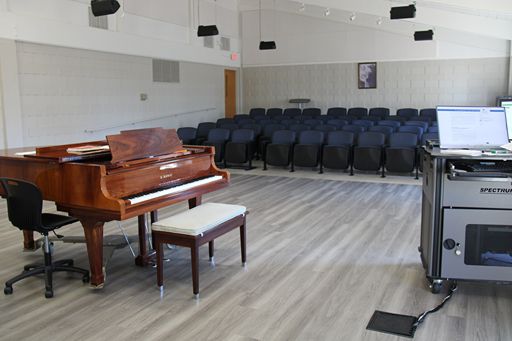 A close-up view of the performance area in the Dora Jane Ledgerwood Ellis Recital Hall featuring a polished wood grand piano and a mobile "Spectrum" professional AV equipment cart. The background shows the transition from the wooden stage floor to the tiered seating area.