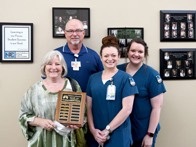 Four people standing in front of a wall. One is holding a plaque and two are in student scrubs.