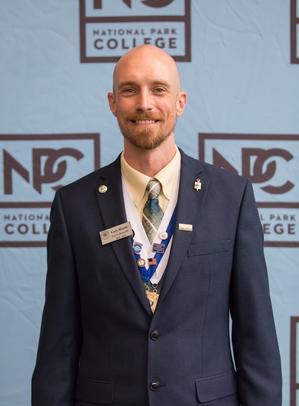 Cory Morris in a suit and tie with his PTK regalia on standing in front of the NPC picture screen.