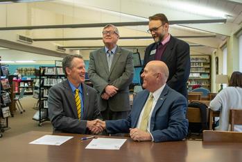 Dr. Hogan and Dr. Berry shaking hands over a contract with two school employees behing them.