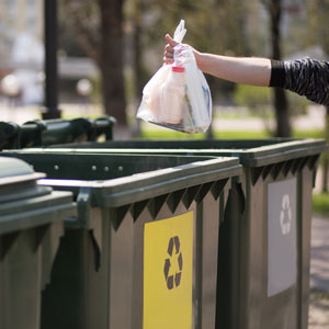 Person holding a trash sack over a garbage can with a recycle sign on front of the can.