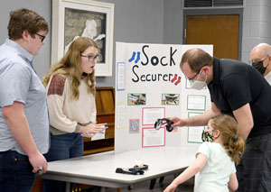 Students with a display board of their project Sock Secure with a man and child holding the prototype.