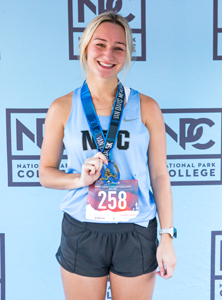 Brooke Wyatt in cross country uniform standing in front of NPC backdrop holding medal.