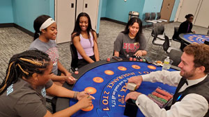 Two casino tables, 5 young women sitting around the table, dealer shuffling deck of cards.
