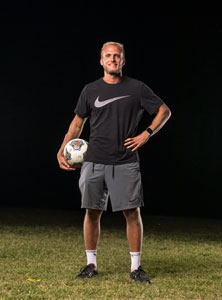 Soccer coach Grant Gartner standing on field with a soccer ball in his arms.