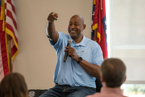 Sidney Moncrief sitting on chair with microphone in one hand pointing to the audience with other hand.
