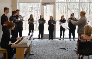 People standing in atrium with music books in hand being led by instructor with a person playing the piano.