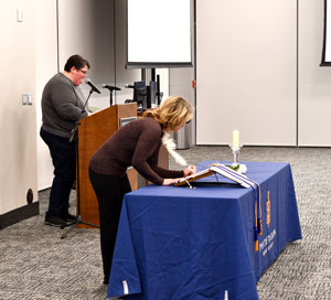 One person standing at a podium another leaned over a table signing a book in a conference room.