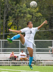 NPC women's division soccer player with a soccer ball coming at her while she jumps in the air, right leg up to kick the ball with arms splayed out to her sides.