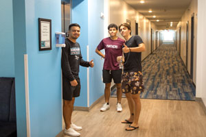 Three men standing in residence hall hallway, two with thumbs up.