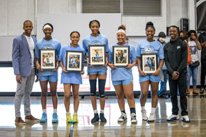 Flanked by coaches, sophomore basketball players stand on court holding pictures of themselves.