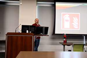 Thomas Gage standing behind a podium with a white screen to the side with a fire extinguisher displayed.