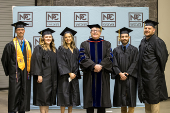 Six people stand in graduation cap and gowns in front of blue background with NPC logo on it.