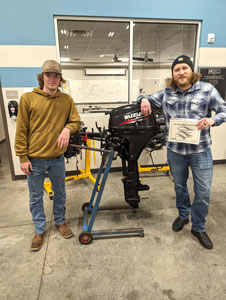 Two students standing in front of boat motor one holding a certificate.