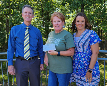 Three people standing in front of trees, the one in the middle holds a check.