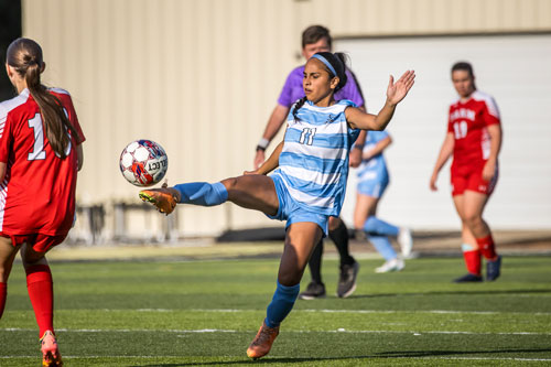 NPC women's soccer team on the field against opponents. NPC player has leg extended, foot stretched about to kick the ball.