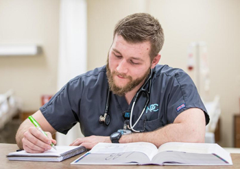 A person reading a textbook taking notes while in scrubs and a stethoscope around their neck.