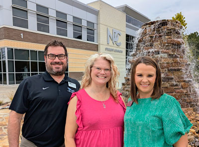 Three people standing in front of a fountain and the NPC Student Commons.