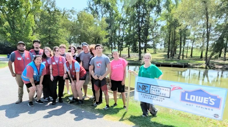 Group of people standing in front of a gody of water with a banner saying National Park College and Lowes.