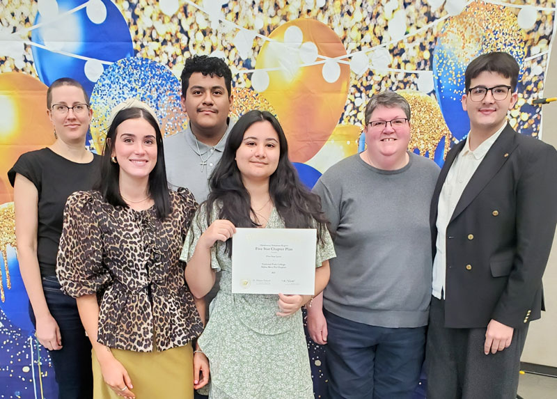 Six people standing in front of a background of balloons and lights. One holding a certificate.