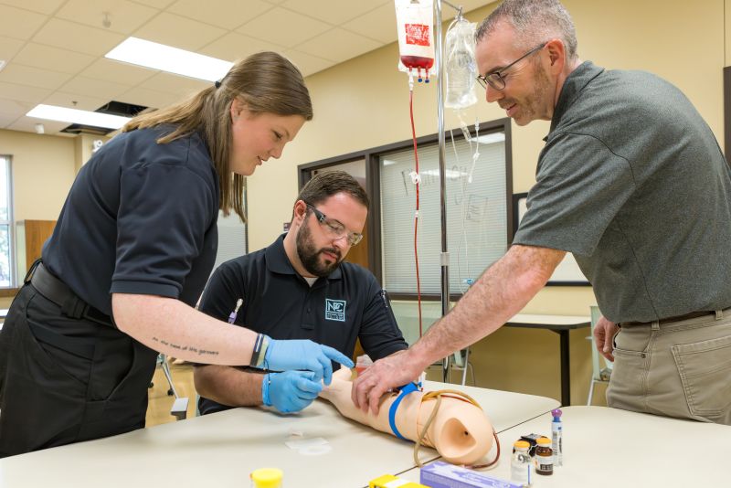 Three people putting an iv in a medical reenactment arm.